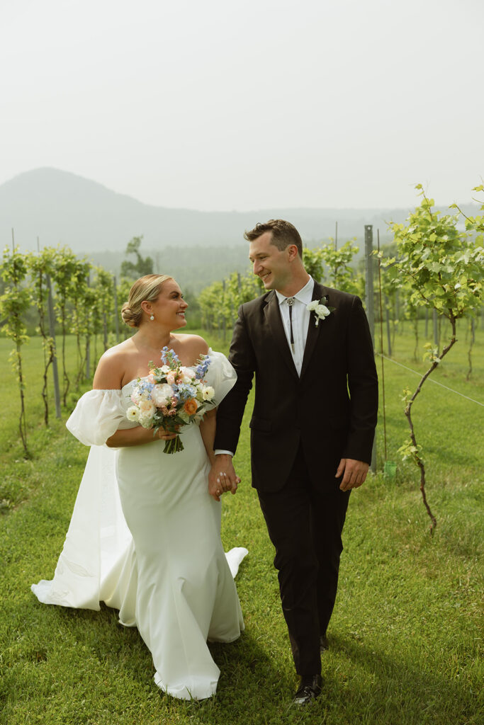 Couple sharing a special look as they walk through the vineyard. Bride is holding a lush pastel bouquet featuring sunshine butter yellow florals. 