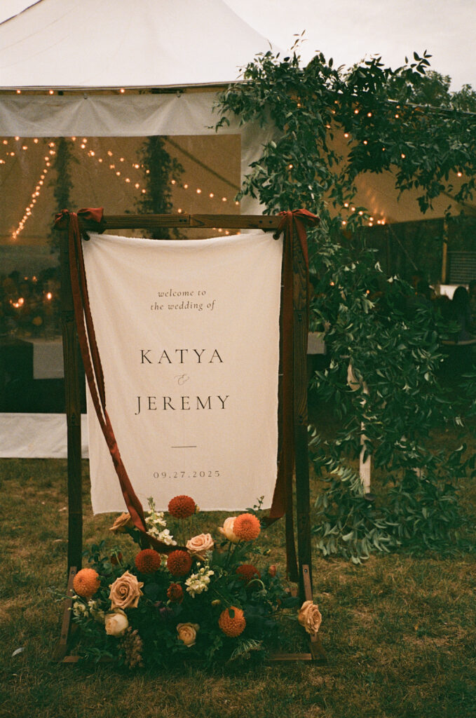 A linen welcome sign flowing in the wind, with florals at the base which welcomes guests to a wedding at Hill Farm Inn. 
