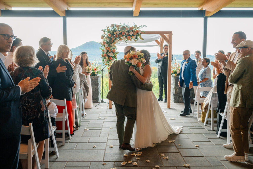 A couple kiss at the end of their aisle with their loved ones clapping next to them. An arbor can be seen in the background decorated with a top corner arrangement. 