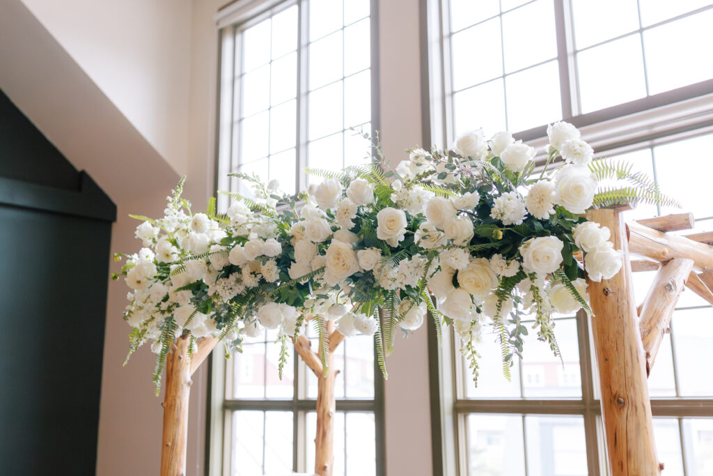 Wedding ceremony featuring all white flowers, ferms and greenery against a wooden arbor. 