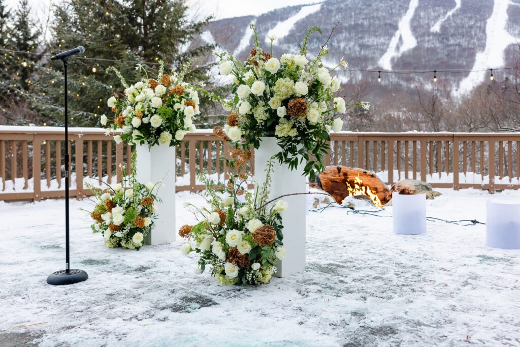 Outdoor floral arrangements featuring two white pillars each with an arrangement sitting on top of the pillar and at the bottom of each.